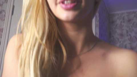 Media: Video of a young woman with long blonde hair, light skin, and full lips. She is topless, wearing a thin necklace. Background features floral-patterned wallpaper and a doorframe.