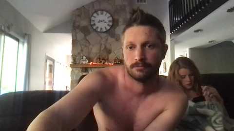 Media: Video of a shirtless man with a beard, dark hair, and fair skin, sitting next to a blonde woman in a striped top, in a modern living room with a stone fireplace, clock, and decorative shelves.