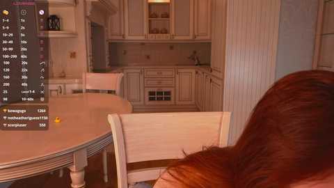 Media: Video of a cozy, modern kitchen with white cabinetry, a round wooden table, and a person with long red hair sitting at the table.