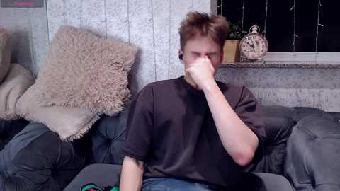 Media: A video of a young man with short, spiky hair wearing a black shirt, sitting on a gray sofa, covering his nose with his right hand, surrounded by textured pillows and a window with string lights.