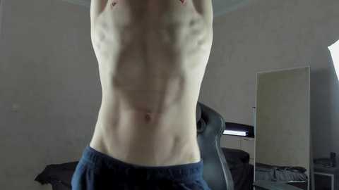 Media: Video of a shirtless, fit, pale-skinned man with defined abs and no visible nipples, wearing blue sweatpants, standing in a dimly lit, cluttered bedroom with a black gaming chair and a large mirror.