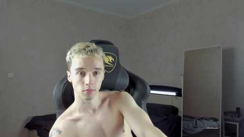 Media: Video of a shirtless, muscular, young man with short blonde hair sitting in a black gaming chair, in a dimly lit room with a plain wall, a mirror, and a nightstand.