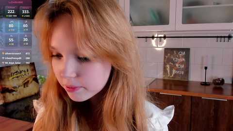 Media: Video of a young Caucasian woman with long, wavy blonde hair in a modern kitchen. She wears a white top, and the background includes white tiles, wooden cabinets, and a lit candle.