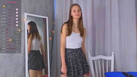 Media: Video of a young Caucasian woman with long, straight brown hair, wearing a white tank top and black floral skirt, standing in a modern bedroom with a white wooden bed, gray wall, and calendar.