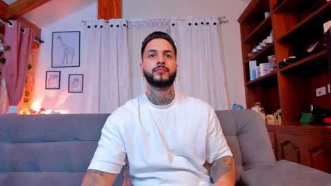 Media: Video of a bearded man in a white T-shirt, sitting on a gray couch, surrounded by wooden bookshelves, framed art, and lit candles in a cozy living room.