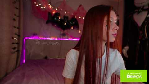 Media: Video of a young woman with long, straight red hair, fair skin, and light makeup, standing in a dimly lit bedroom with a pink bedspread, fairy lights, and a Halloween decoration on the wall.