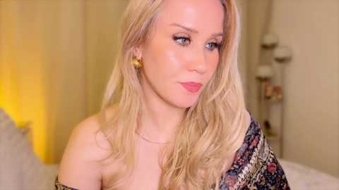 Media: Video of a fair-skinned woman with long, wavy blonde hair, wearing gold earrings and a colorful, patterned shawl. She has a serene expression, set against a softly lit, beige-toned background.