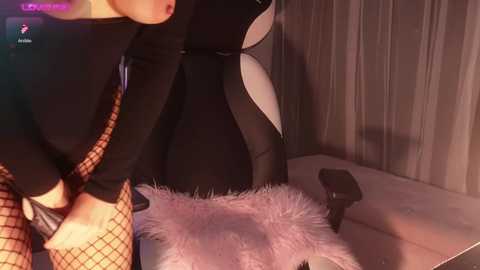 Media: Video of a woman in black fishnet stockings and a black bodysuit, leaning over a white fur chair in a dimly lit room with gray curtains.