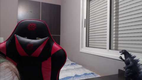 Media: A video featuring a modern gaming chair with red accents and a plush texture, next to a white window with horizontal blinds, and a small Godzilla figurine on a white and blue rug.