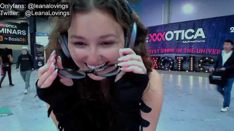 Media: Video of a smiling, dark-haired woman in black attire with headphones, standing in a dimly lit, modern room. Background includes a \"Exotic\" banner and social media handles.