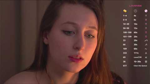 Media: Video of a young woman with fair skin, green eyes, and long brown hair, wearing a black top, in a dimly lit room.