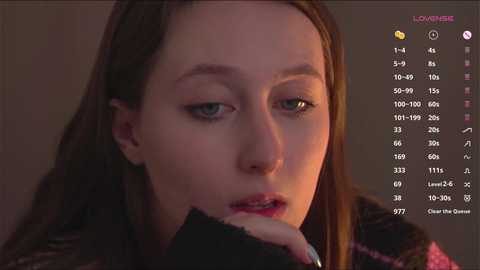 Media: A close-up video of a young woman with fair skin, green eyes, and straight brown hair, wearing a black jacket, appearing contemplative with a soft, warm background.