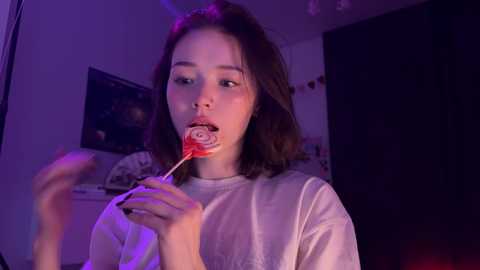 Media: Video of a young woman with shoulder-length brown hair eating a lollipop in a dimly lit room, wearing a white T-shirt.