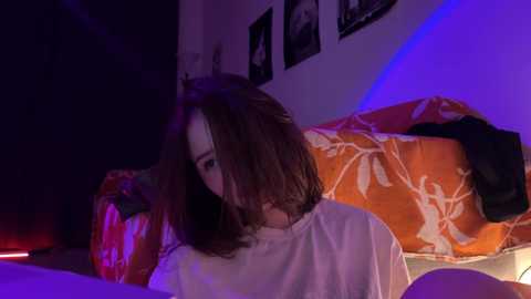 Media: Video of a young woman with shoulder-length brown hair, wearing a white t-shirt, lying on an orange floral bedspread in a dimly lit room with purple lighting.