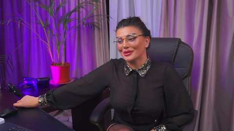 Media: Video of a middle-aged woman with a medium build, wearing glasses and a black blouse with a necklace, sitting at a desk with a phone and plant in the background.