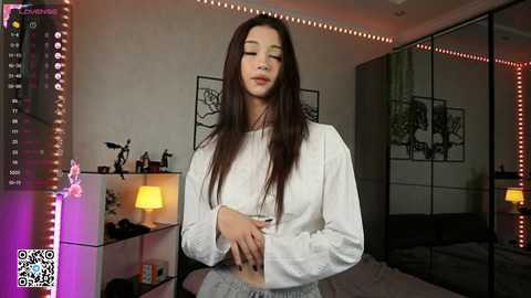 Media: Video of an Asian woman with long black hair, wearing a white crop top and light gray pants, standing in a dimly lit bedroom with a bed, mirror, and decorative shelves.