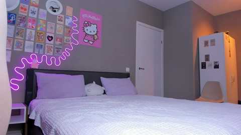 Media: Video of a cozy, minimalist bedroom with a gray wall adorned with Hello Kitty posters, a neatly made bed with lavender linens, and a white fridge decorated with photos.