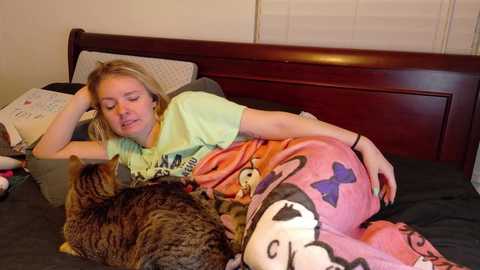 Media: Video of a blonde woman with fair skin, wearing a green shirt and pink pajama bottoms, lying on a dark bed with a gray blanket. Next to her, a striped tabby cat sleeps peacefully.