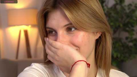 Media: Video of a young woman with light brown hair, freckles, and a red bracelet, hiding her face with her hand, smiling. Background shows a lamp, beige wall, and green plant.