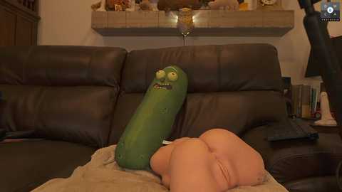 Media: A video of a dark brown leather couch with a large, green, pickle-shaped plush toy sitting upright, wearing a small hat and glasses, and a small person's bare buttocks in the foreground.