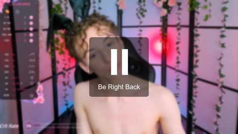 Media: Video of a shirtless, young man with curly brown hair in a dimly lit room with hanging plants and pinkish-purple lighting. Text overlay reads \"Be Right Back.\