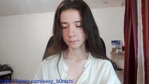 Media: A young woman with fair skin, straight dark hair, and a slim build, wearing a white robe, sits in a dimly lit room with a white wall, a red curtain, and a cluttered shelf in the background.