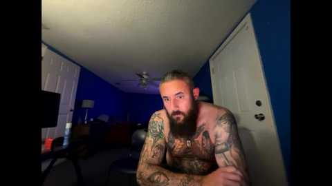 Media: A video of a muscular, bearded, heavily tattooed man with short, light brown hair, shirtless, leaning on a desk in a dimly-lit room with blue walls and white doors.