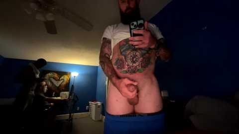 Media: A video of a bearded man with tattoos, including a large chest tattoo, holding a phone to take a mirror selfie. He's in a dimly lit room, standing with his blue pants pulled down, exposing his flaccid penis.