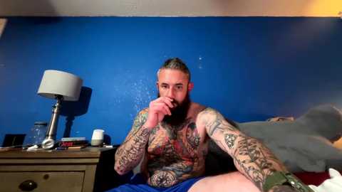 Media: Video of a muscular, tattooed man with a beard, sitting shirtless on a bed, blue wall, nightstand with lamp, and books.