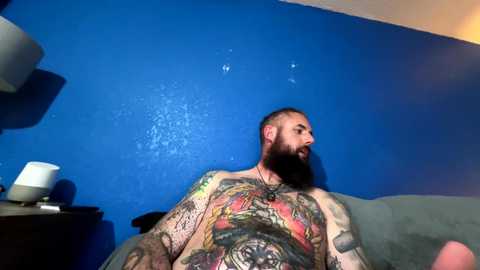 Media: Video of a tattooed, bearded man with dark skin and a muscular build, sitting on a blue couch against a blue wall. He wears a black necklace and a black shirt, showcasing intricate tattoos.