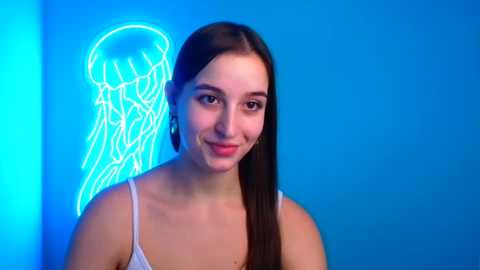 Media: A video of a young woman with straight, dark hair and light skin, smiling, wearing a white tank top, standing in front of a glowing blue jellyfish neon sign against a solid blue background.