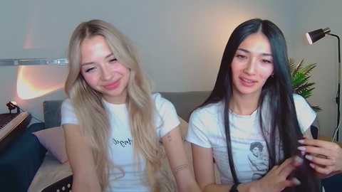 Media: Video of two young women with long blonde and black hair, smiling, wearing white t-shirts with text, sitting on a couch in a brightly lit room.