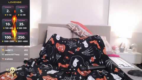 Media: A video of a messy bed with a black sheet adorned with Halloween-themed orange pumpkins and bats. The background shows a modern, minimalist bedroom with white walls and a bedside table holding a lamp.