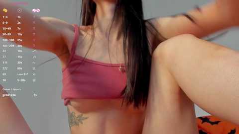 Media: Video of a woman with long black hair, wearing a pink crop top, revealing a tattoo on her side, sitting with legs apart, in a dimly lit room.