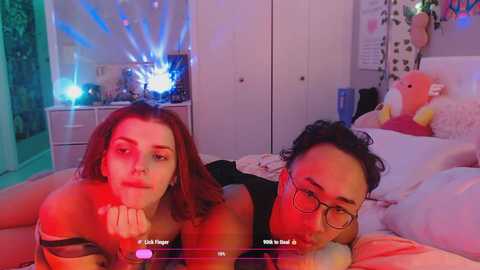 Media: A video of two women, one with red hair and the other with curly black hair, lying on a bed. Both have red makeup on their faces. The room is lit with blue and red lights.