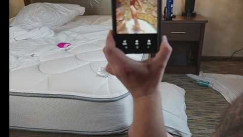 Media: Video of a person taking a selfie in a hotel room, capturing an unmade bed with white sheets, a pink vibrator on the bed, and a nightstand with a lamp and toiletries.