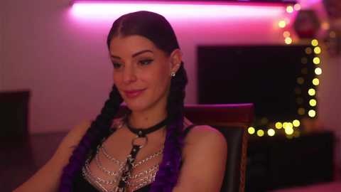 Media: Video of a woman with dark hair in braids, wearing a black leather choker with purple accents, and a sheer, sequined top, seated in a dimly lit room with neon lights and bokeh.