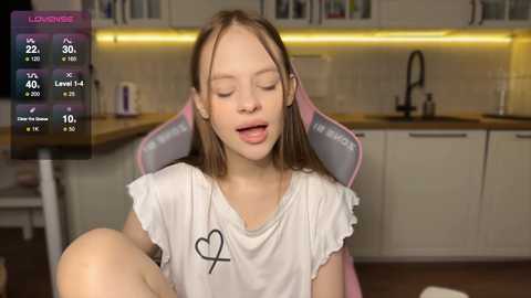 Media: A video of a young girl with light skin and long brown hair, wearing a white T-shirt with a heart graphic, sitting in a pink gaming chair in a modern kitchen.