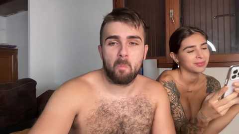 Media: Video of a shirtless man with a full beard, hairy chest, and tattoos, sitting next to a woman with dark hair and tattoos, both looking at a smartphone.