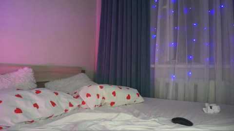 Media: Video of a dimly lit bedroom with a white bed, white pillows, and a duvet covered in red heart patterns. A window with sheer curtains and blue fairy lights casts a soft glow. A black remote control is on the bed.