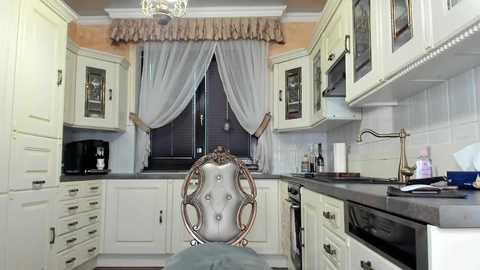 Media: Video of a luxurious, cream-colored kitchen with white cabinets, a high-back chair with ornate detailing, and a window with sheer white drapes.