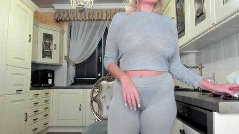 Media: Video of a middle-aged woman with blonde hair in a light blue, long-sleeved crop top and matching leggings, standing in a modern kitchen with white cabinets and a dark countertop.