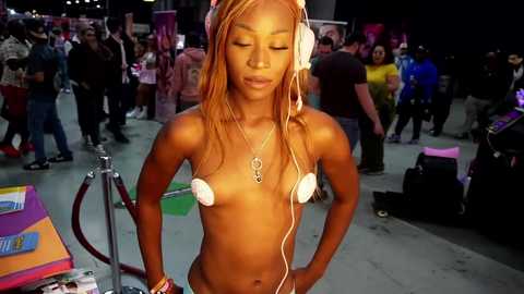 Media: Video of a topless, light-skinned Black woman with long, straight orange hair, wearing white headphones and nipple covers, standing in a crowded outdoor festival setting.