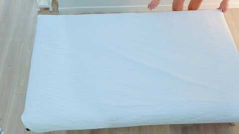 Media: Video of a woman with fair skin, wearing a white bra, pulling a white, textured mattress onto a light wooden floor. The background includes a white door and a small, partially visible white dresser.