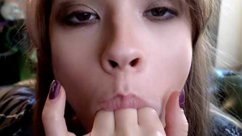 Media: Video of a close-up of a Caucasian woman with fair skin, light brown hair, and dark brown eyes, licking her fingers with purple nail polish. She wears a dark-colored jacket.