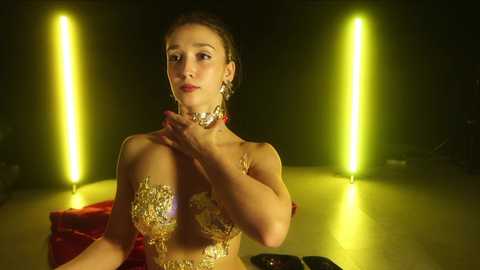 Media: Video of a fair-skinned woman with brown hair in a braided updo, wearing elaborate, gold-sequined, sheer, midriff-baring attire, set against a dimly lit, studio background with glowing, vertical neon lights.