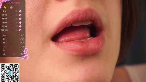 Media: A close-up video of a woman's open mouth, showcasing her full lips and tongue. The background is a blurred, dark room. A QR code and a pink, floral pattern are visible in the upper left corner.