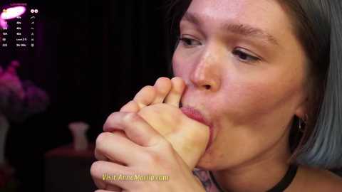 Media: A video of an older woman with gray hair licking a banana, wearing a floral top, in a dimly lit room. The background is blurred.