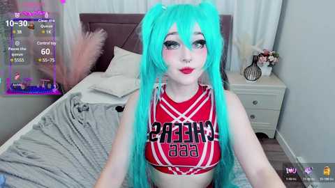 Media: Video of a pale-skinned, green-haired woman with pink makeup, wearing a red \"Greenhouse\" crop top, sitting on a bed in a modern bedroom.