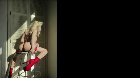 Media: Video of a blonde woman in red lingerie and boots, sitting provocatively on a stool in a dimly lit room, with shadow play creating dramatic contrast.